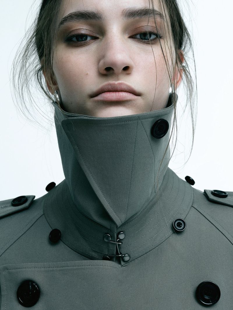 Close up portrait of a model wearing a high collared grey trench coat with black buttons against a plain white background