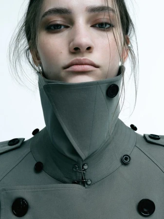 Close up portrait of a model wearing a high collared grey trench coat with black buttons against a plain white background