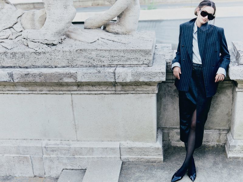 Model in dark sunglasses wearing a pinstripe blazer and dark skirt leaning against ornate stone architecture