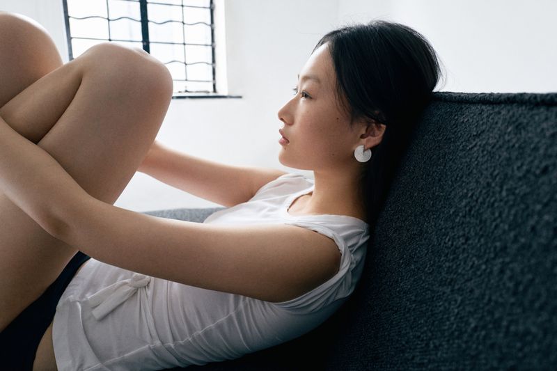 Model seated sideways on a dark textured sofa wearing a white top and round earrings against a bright interior wall.
