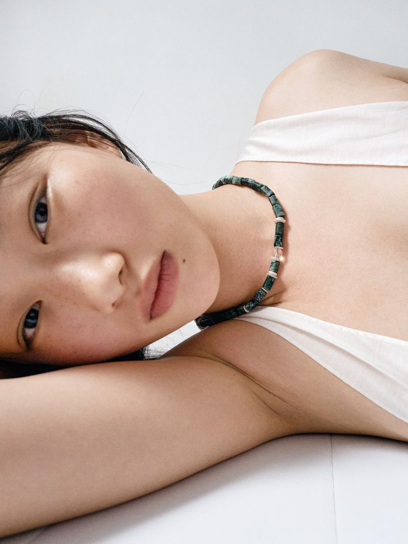 Close-up portrait of model Zhao Ziqi wearing a dark green beaded necklace and white top against a clean white background.
