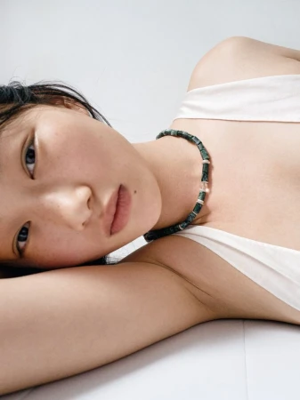Close-up portrait of model Zhao Ziqi wearing a dark green beaded necklace and white top against a clean white background.