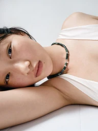 Close-up portrait of model Zhao Ziqi wearing a dark green beaded necklace and white top against a clean white background.