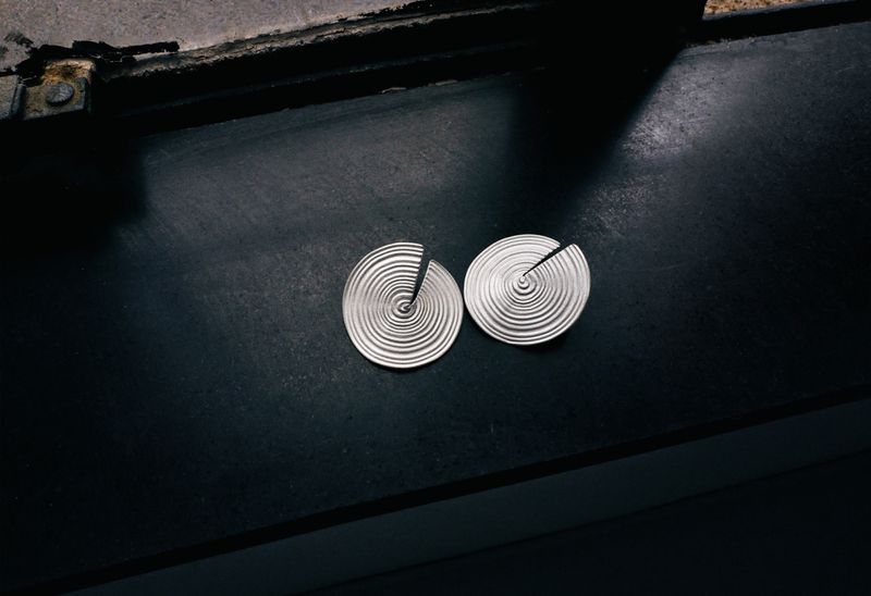 Two circular silver earrings featuring concentric spiral ridges resting on a dark matte surface