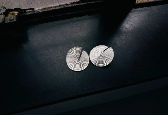 Two circular silver earrings featuring concentric spiral ridges resting on a dark matte surface