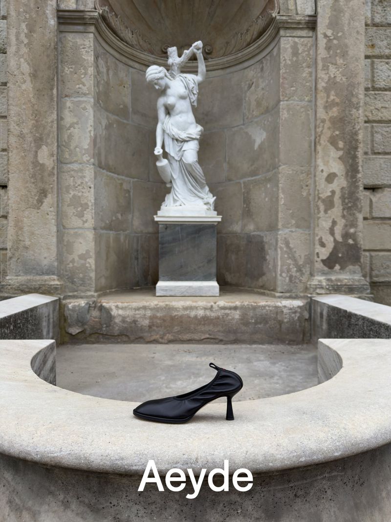 Black heeled shoe resting on a stone fountain ledge with a classical white statue in the background for Aeyde Pre Fall 26