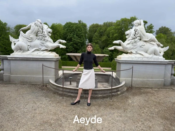Model standing outdoors between large stone lion statues and a fountain for Aeyde Pre Fall 26 campaign