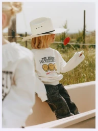 Child wearing a white cowboy hat and smiley face sweatshirt holds a red poppy flower outdoors in the project The Journey.