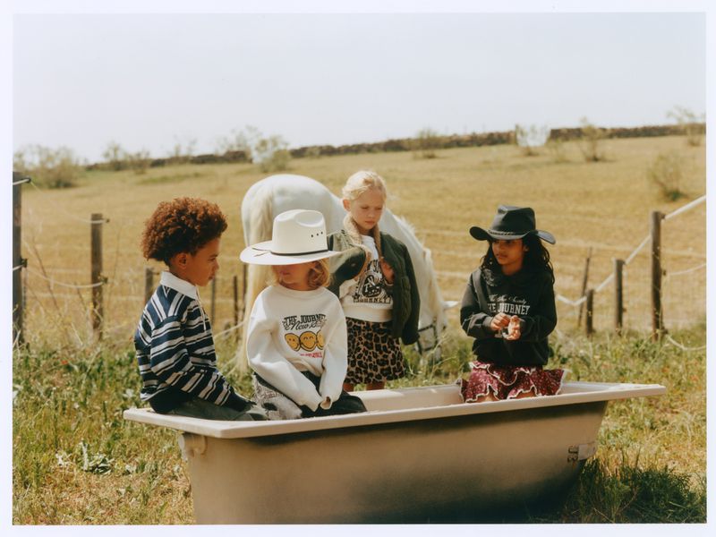 Four children in casual clothing posed together around a vintage bathtub in an open field with a white horse standing behind them.