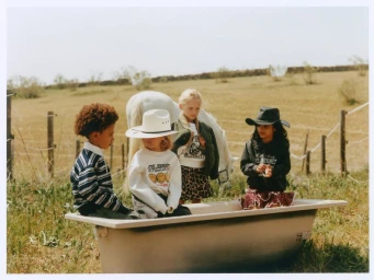 Four children in casual clothing posed together around a vintage bathtub in an open field with a white horse standing behind them.