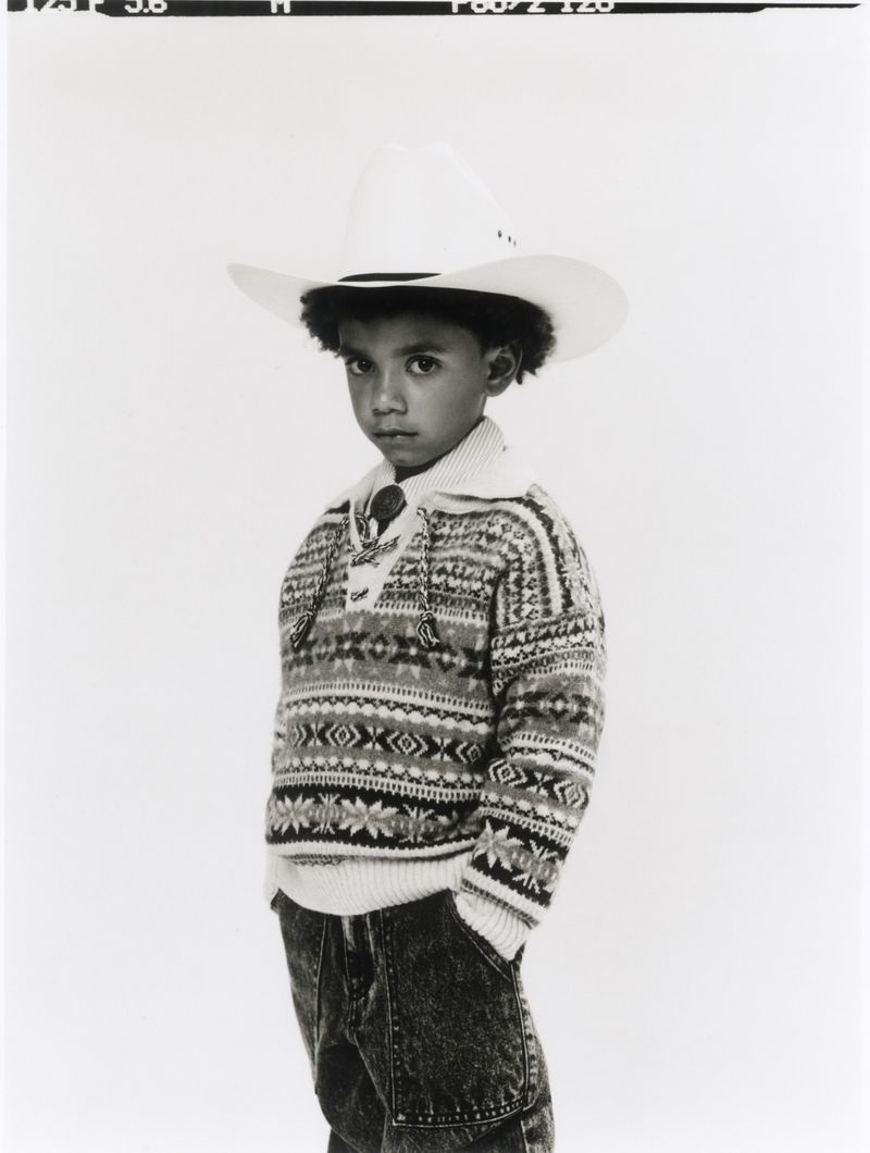 Young child wearing a white cowboy hat and patterned sweater against a neutral background.