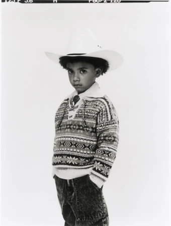 Young child wearing a white cowboy hat and patterned sweater against a neutral background.