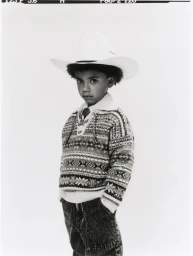 Young child wearing a white cowboy hat and patterned sweater against a neutral background.
