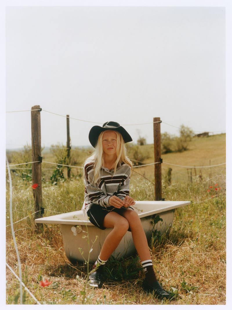 Young blond person wearing a dark wide-brim hat and striped shirt, seated on a vintage bathtub in a grassy field.