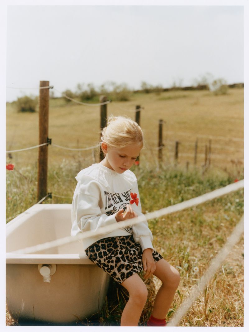 Young child with blonde hair sitting on the edge of an outdoor bathtub in a grassy field wearing a white sweatshirt and leopard print skirt