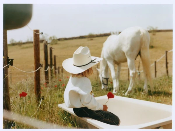 Child wearing a cowboy hat sitting on the edge of an outdoor tub holding a red flower while a white horse grazes in a pasture.