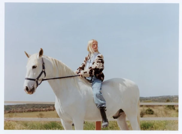 Blonde woman riding a white horse in an outdoor field wearing a cow print jacket and blue jeans