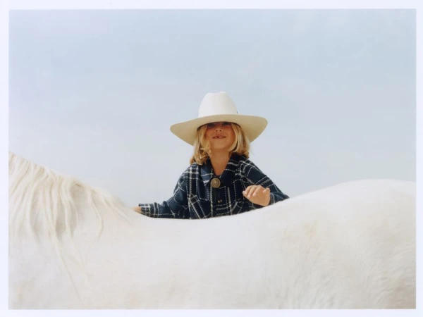 Young child wearing a white cowboy hat and plaid shirt sitting behind a white horse against a pale sky.