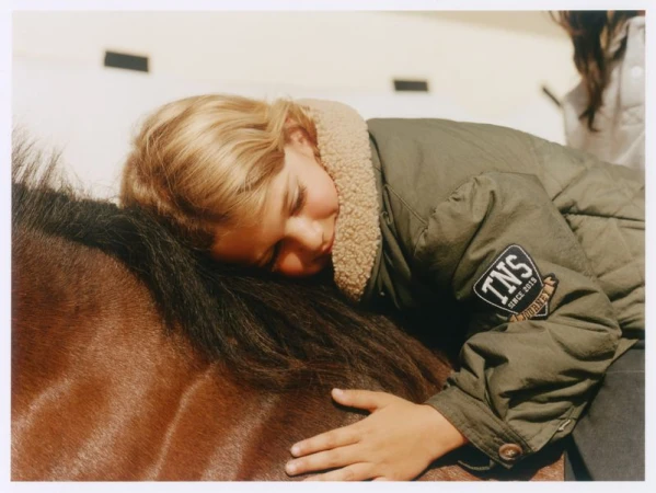 Blonde child resting head against a brown horse wearing a green jacket with a patch