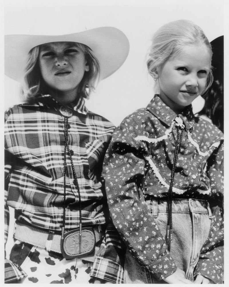 Black and white portrait of two young children wearing western shirts and bolo ties, captured outdoors in bright sunlight.