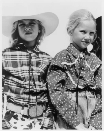 Black and white portrait of two young children wearing western shirts and bolo ties, captured outdoors in bright sunlight.