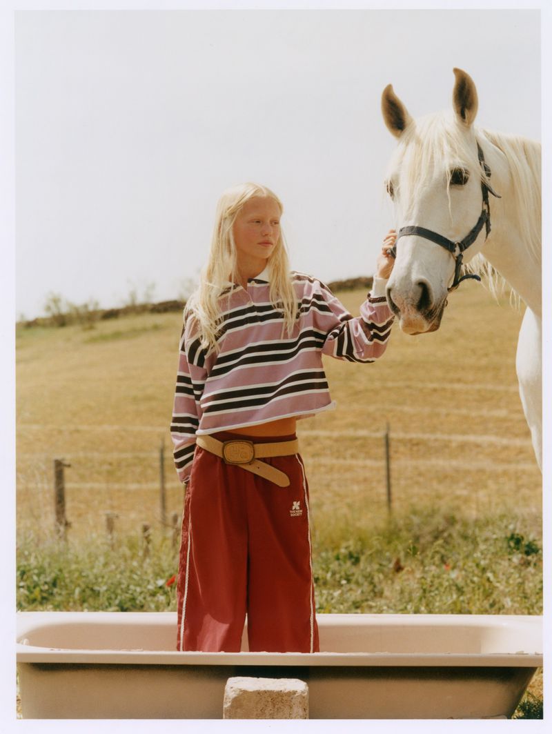 Blonde female model standing in a tub outdoors holding the reins of a white horse in a meadow