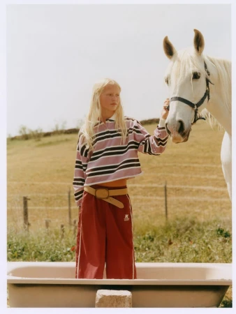 Blonde female model standing in a tub outdoors holding the reins of a white horse in a meadow