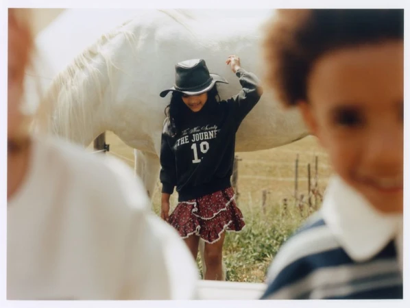 Child wearing a cowboy hat and black graphic sweatshirt posing outdoors near a white horse with other children in the blurred foreground.