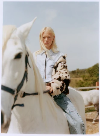 Blonde young woman riding a white horse wearing a cow-print jacket and striped shirt