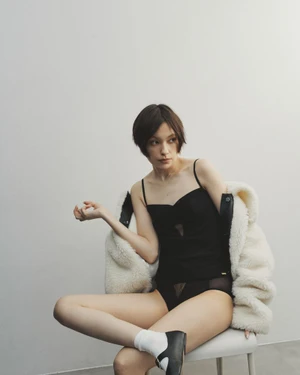 Model with short dark hair wearing a black camisole and white fluffy jacket seated on a white chair against a plain white background.
