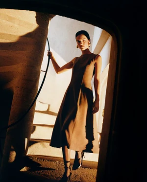 Model standing on stone steps in a structured brown dress, captured from a dark architectural frame with dramatic warm lighting.