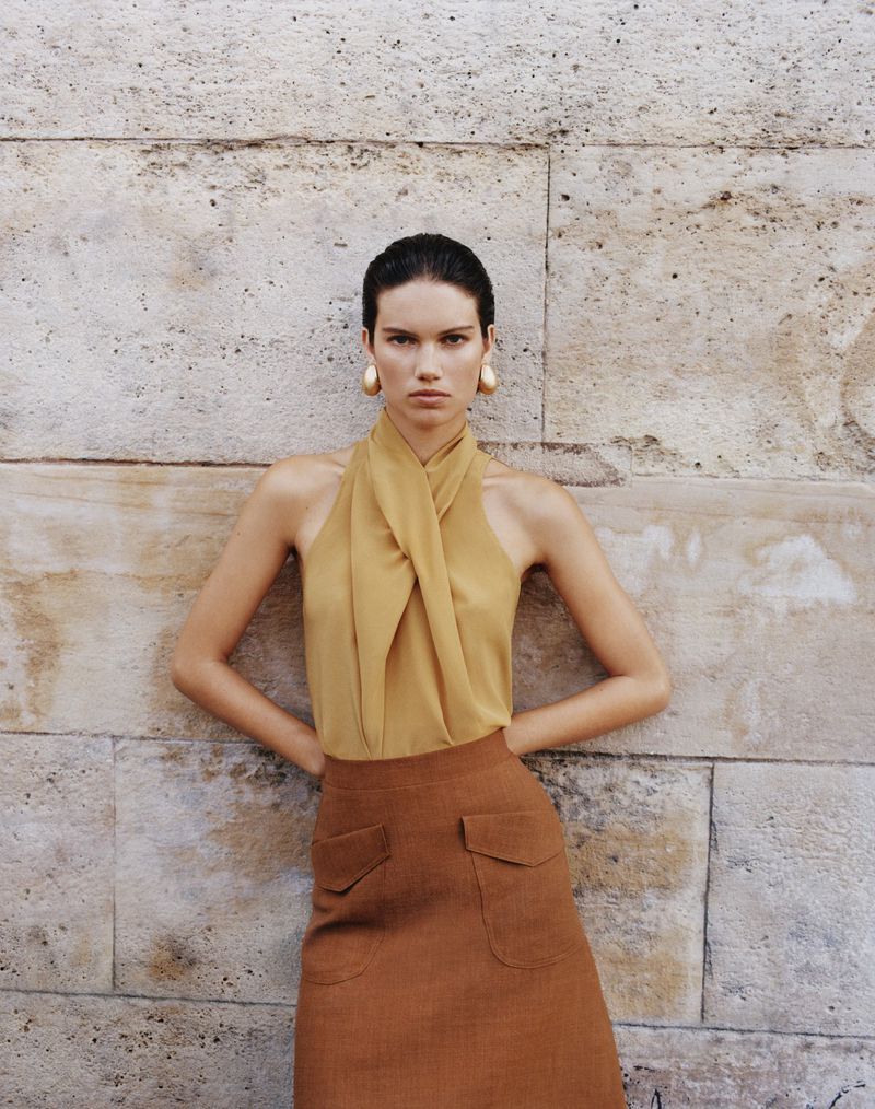Model in a mustard halter top and brown skirt standing against a textured stone wall