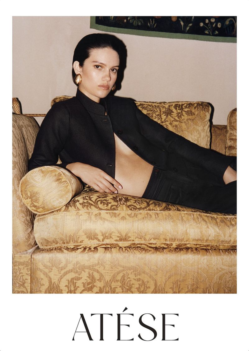 Model reclining on an ornate patterned sofa wearing a dark jacket and gold earrings in the At©se campaign
