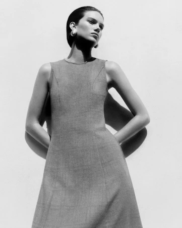 Model in a grey sleeveless dress posing against a plain white wall with harsh shadows cast behind her.