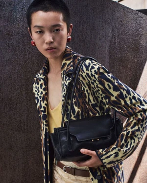 Model with short dark hair wearing a leopard print jacket and holding a black leather shoulder bag outdoors