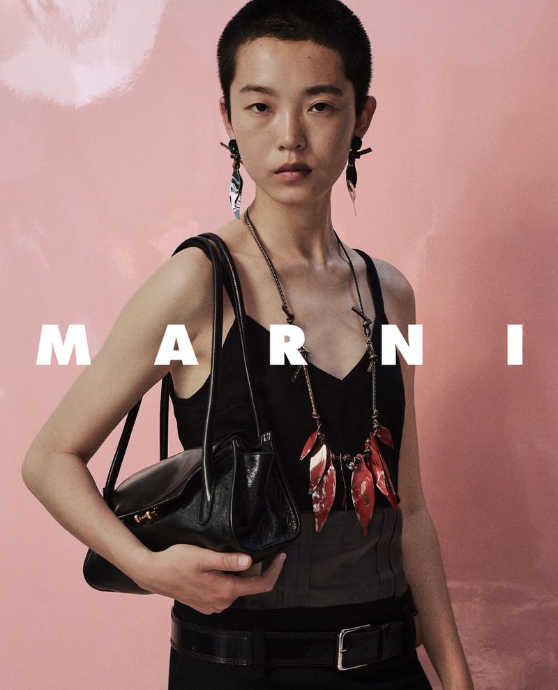 Model with short hair wearing a black top and holding a black leather handbag against a pink background