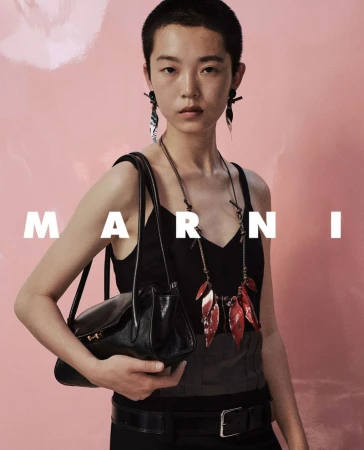 Model with short hair wearing a black top and holding a black leather handbag against a pink background
