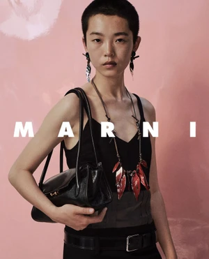 Model with short hair wearing a black top and holding a black leather handbag against a pink background
