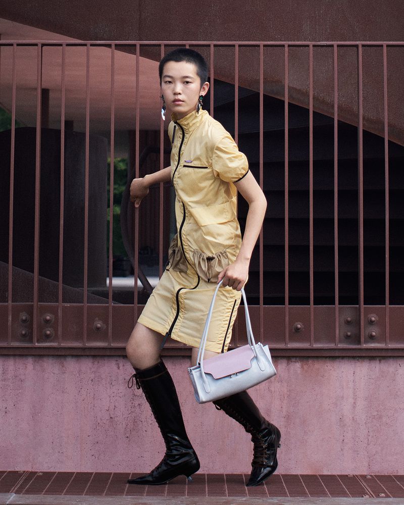 Model in yellow dress and black boots holding a handbag posing behind a metal railing