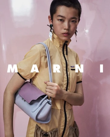 Model with short dark hair wearing a yellow zip-up top holding a light blue and purple shoulder bag against a pink background