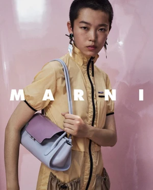 Model with short dark hair wearing a yellow zip-up top holding a light blue and purple shoulder bag against a pink background