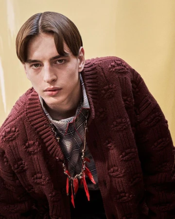 Close-up portrait of a male model wearing a textured burgundy cardigan and plaid shirt against a yellow background.
