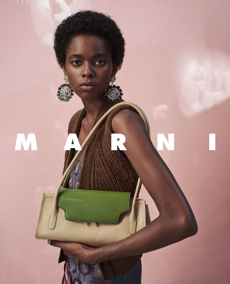 Model with short dark hair wearing ornate earrings and a brown knitted vest holding a green and beige handbag for Marni F/W 2026.