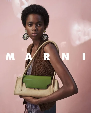 Model with short dark hair wearing ornate earrings and a brown knitted vest holding a green and beige handbag for Marni F/W 2026.