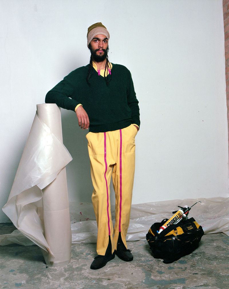 Bearded adult male fashion model in dark green sweater, yellow pants, and head covering posing near roll of plastic.