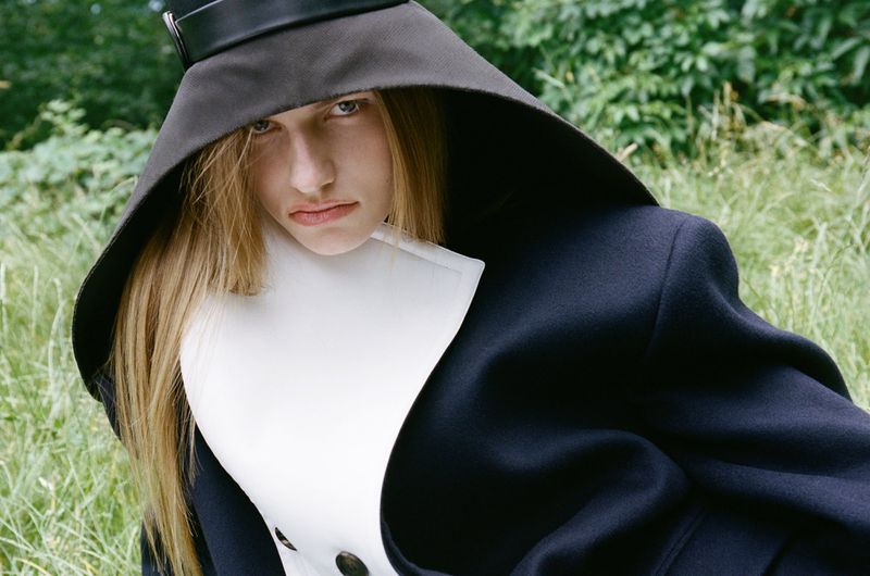 Model Anne Floor Wetemans wearing a dark hooded hat and structured coat outdoors in a green meadow for Harper's Bazaar Italia.