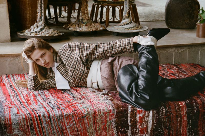Model lying on a patterned red rug wearing a plaid blazer, white wide belt, and black boots for an editorial shoot.