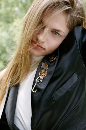 Close-up portrait of a model with long blonde hair wearing a black jacket adorned with pins and medals in an outdoor setting