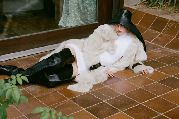 Model lying on brown tiled floor wearing white mini dress, fur coat, tall black boots and wide-brim hat