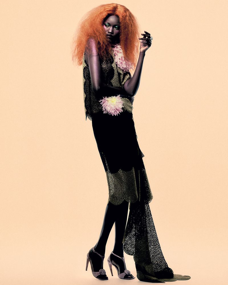 Model Caren Jepkemei with vibrant orange hair wearing a dark patterned dress with feather accents against a plain neutral background.